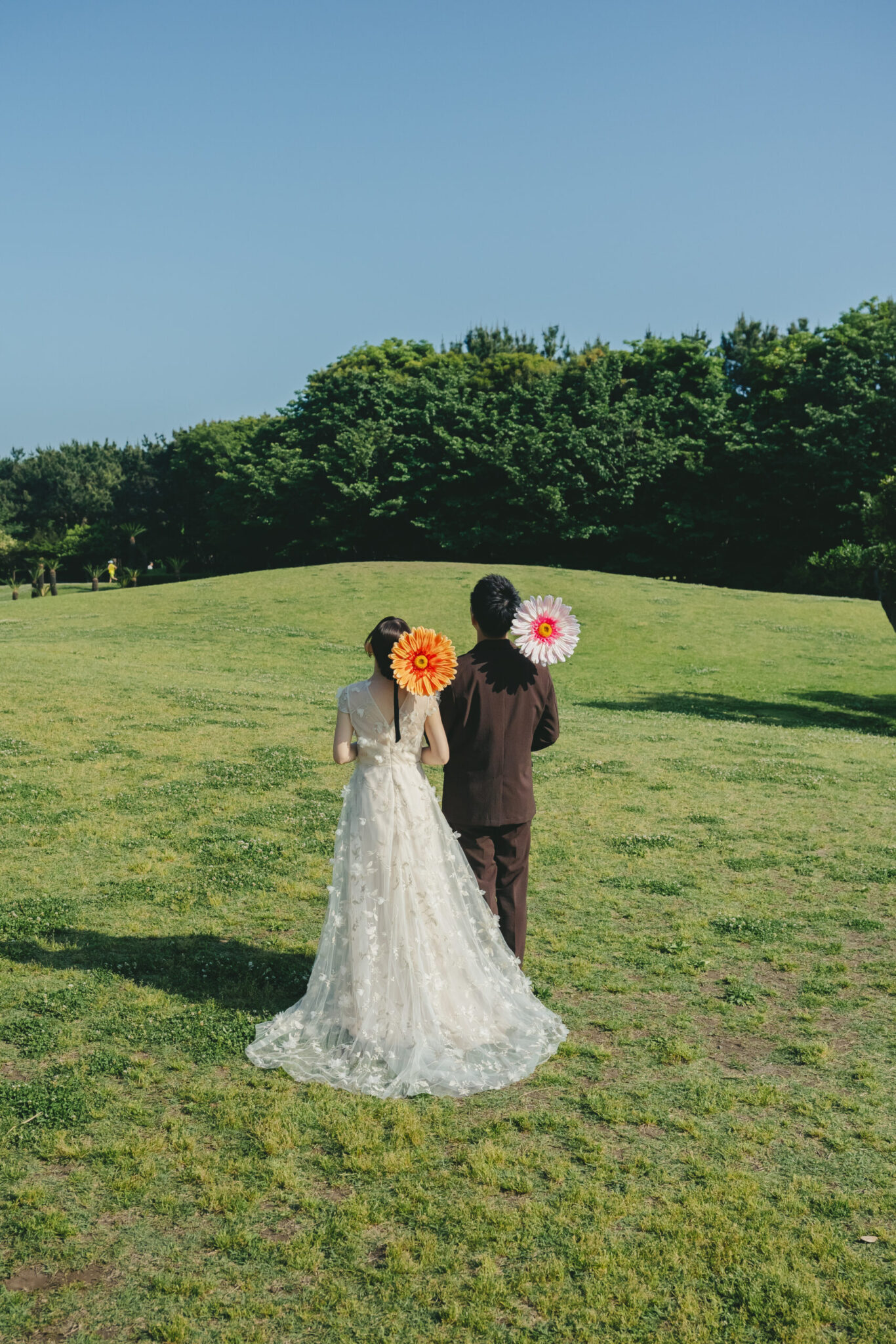 神奈川の辻堂海浜公園の広大な緑の芝生の上で可愛いお花のプロップスを背中に持って佇む新郎新婦の後ろ姿カット
