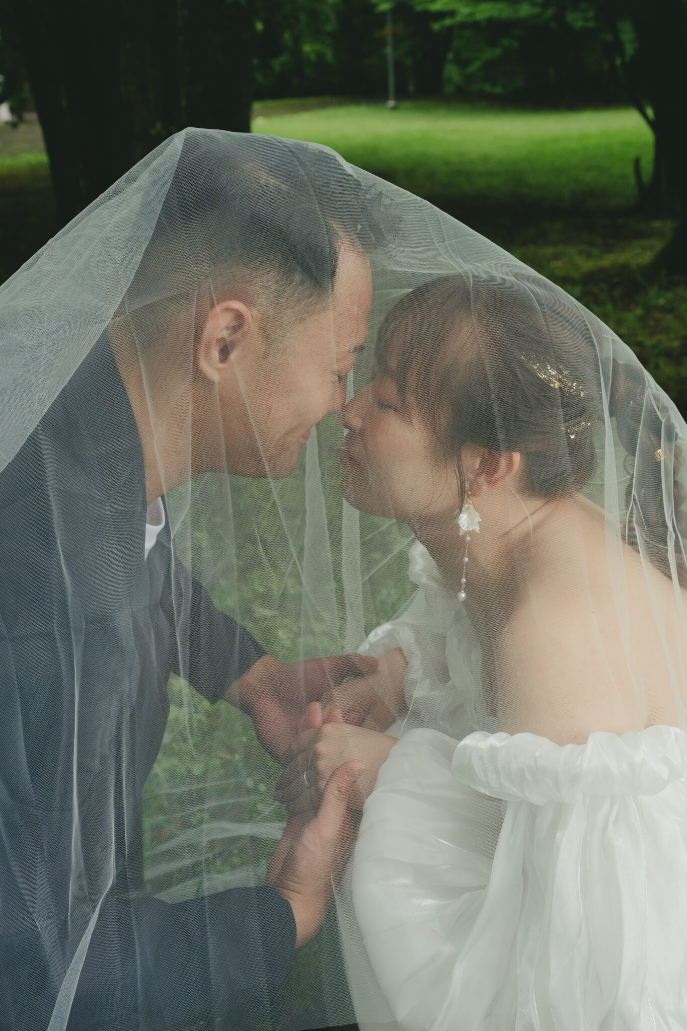 うつのみや文化の森の緑を背景にロングベールを被った新郎新婦の幻想的なウェディングフォト
