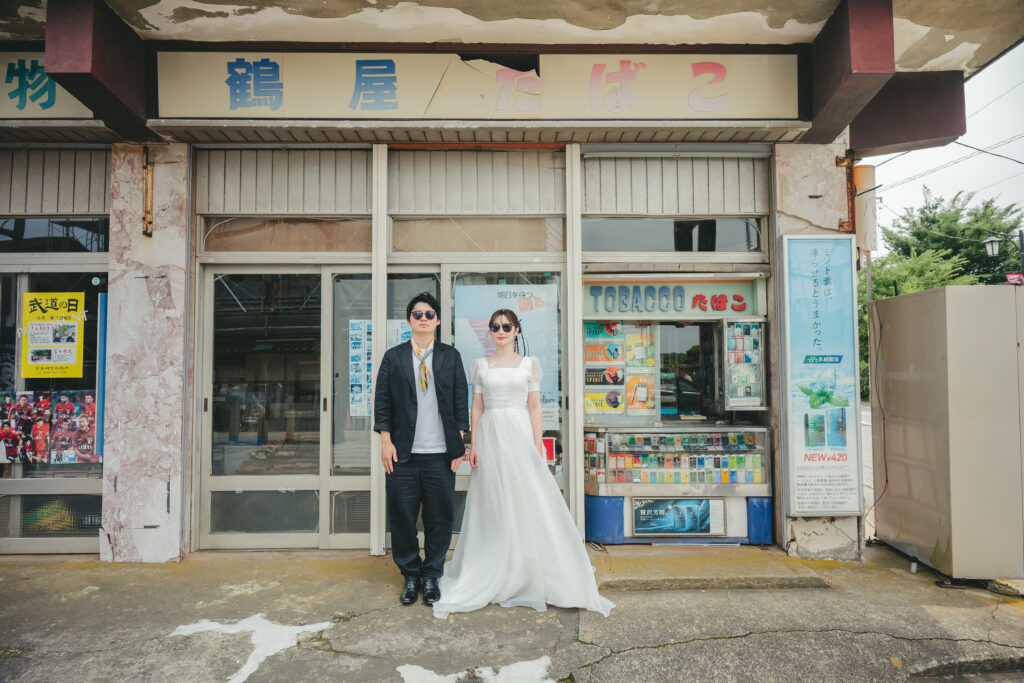 この記事のアイキャッチ画像ですね！昭和レトロなたばこ屋さん（商店）の前でサングラスをかけてビシッと決めている、めちゃくちゃセンスが光る最高の一枚です！ この記事全体の顔になるアイキャッチ画像なので、狙いたい超重要SEOキーワード（茨城 前撮り 鹿島 ロケーションフォト）を網羅した代替テキスト（altテキスト）をご提案します。 コピーして貼り付けてください！ 📝 鹿嶋記事のアイキャッチ用：代替テキスト案 茨城県鹿嶋市のレトロな商店街でタキシードとドレスにサングラスを合わせて並ぶお洒落なロケーション前撮り