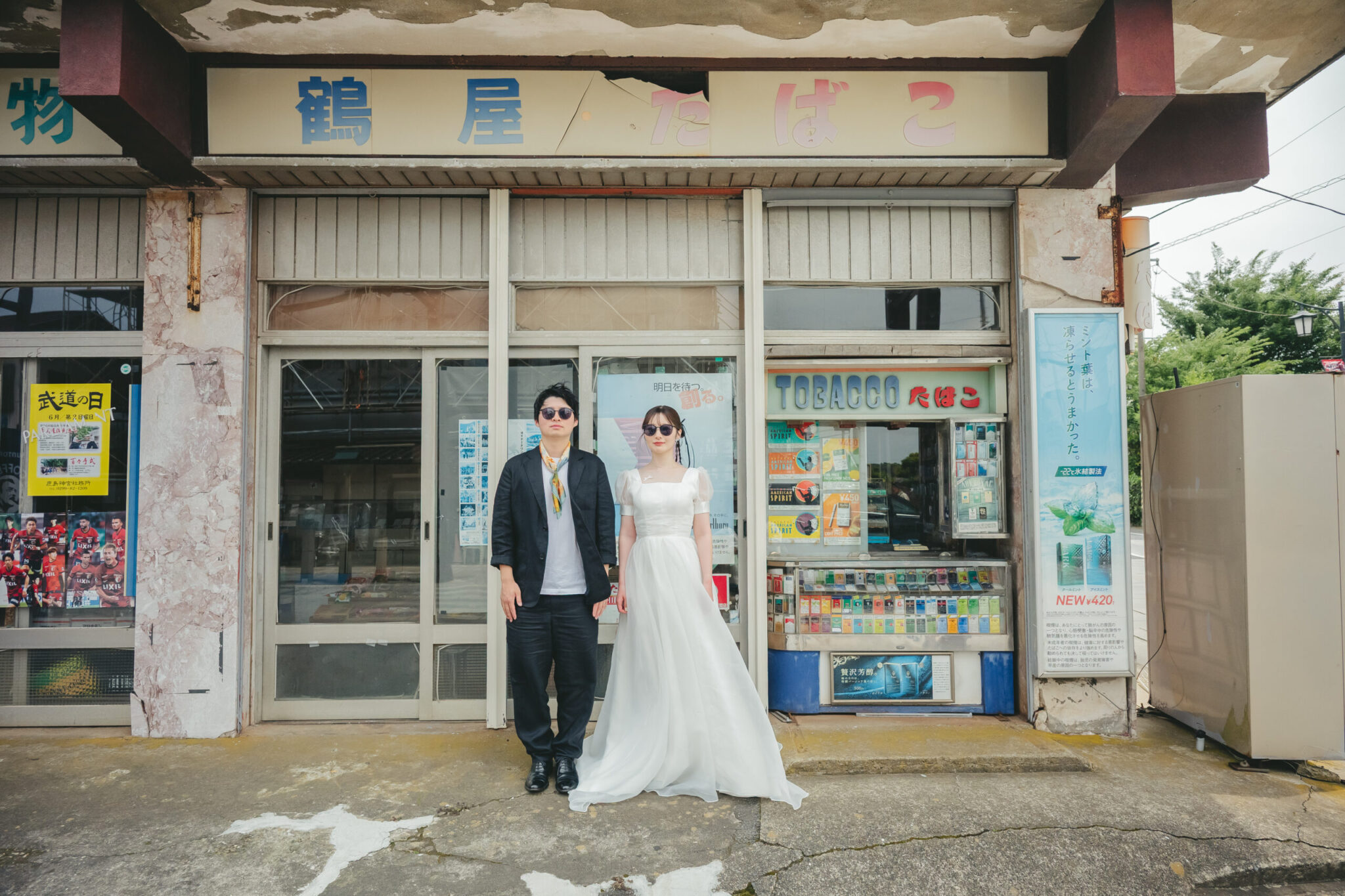 この記事のアイキャッチ画像ですね!昭和レトロなたばこ屋さん(商店)の前でサングラスをかけてビシッと決めている、めちゃくちゃセンスが光る最高の一枚です! この記事全体の顔になるアイキャッチ画像なので、狙いたい超重要SEOキーワード(茨城 前撮り 鹿島 ロケーションフォト)を網羅した代替テキスト(altテキスト)をご提案します。 コピーして貼り付けてください! 📝 鹿嶋記事のアイキャッチ用:代替テキスト案 茨城県鹿嶋市のレトロな商店街でタキシードとドレスにサングラスを合わせて並ぶお洒落なロケーション前撮り