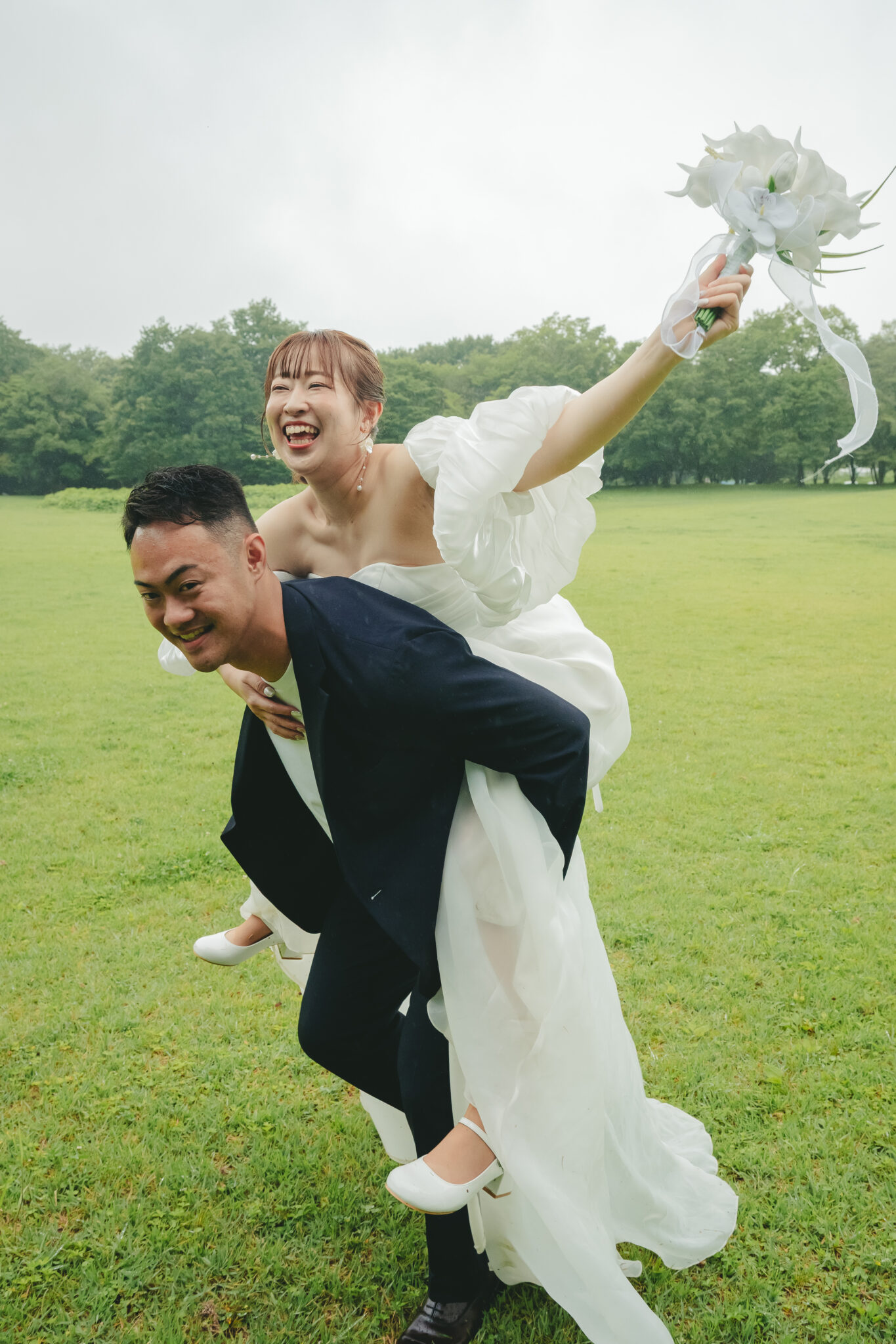 ドレスとタキシード姿で新郎が新婦をおんぶしてるうつのみや文化の森での楽しい前撮り