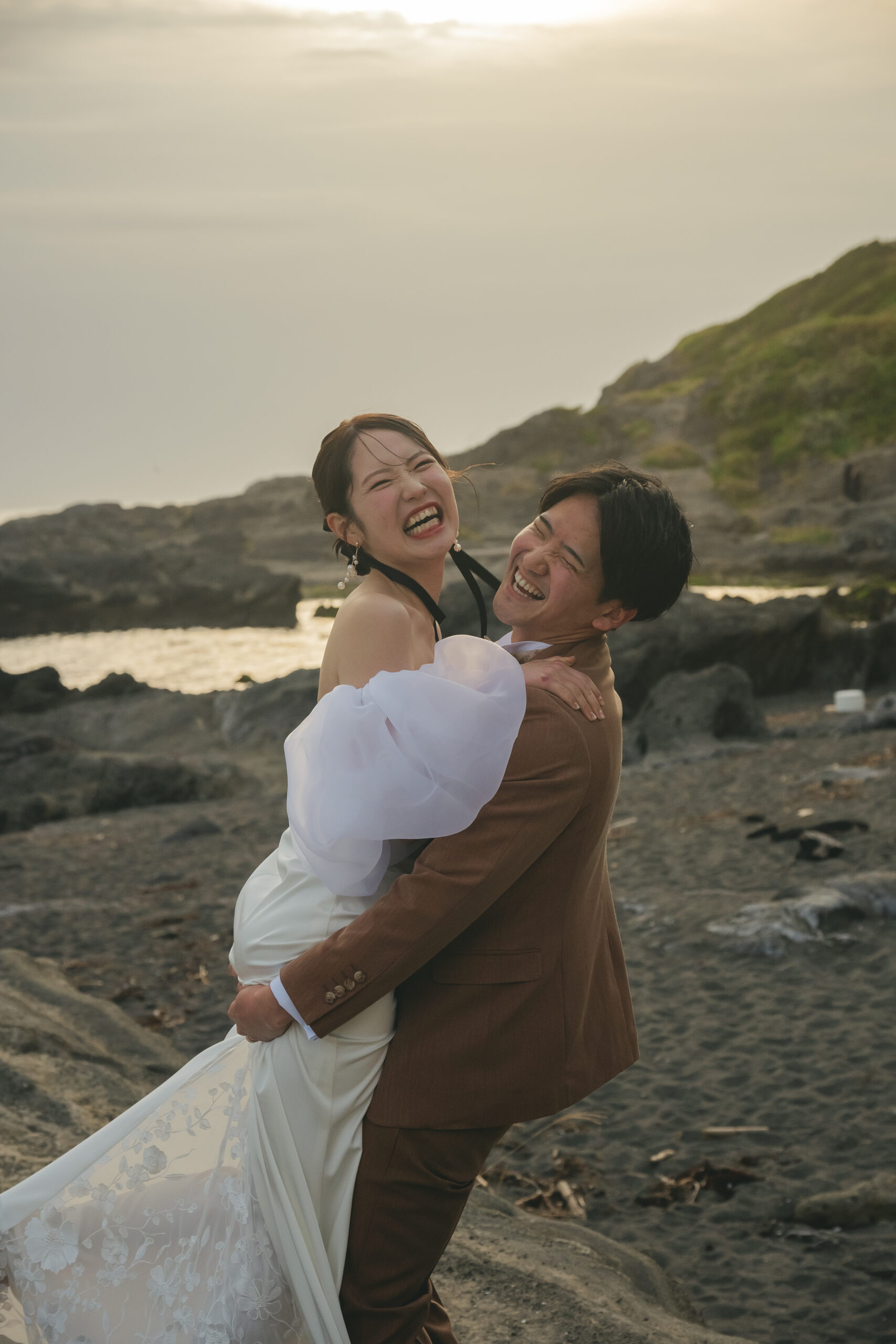 城ヶ島の夕刻の海と岩場を背景に新婦を抱き上げるロマンチックな前撮りポーズ