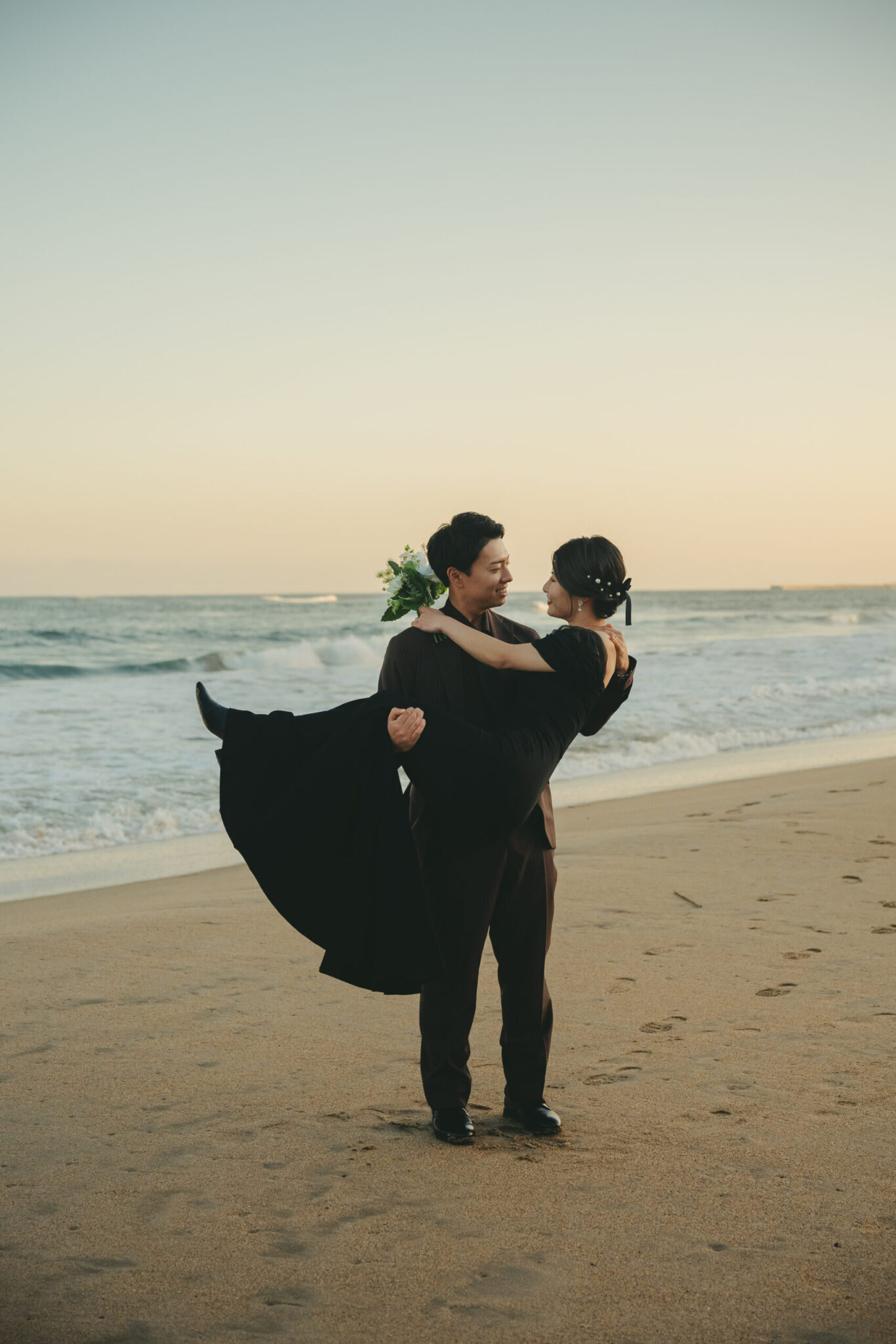 夕暮れ時の久慈浜海水浴場でシックなブラックドレス姿の新婦をお姫様抱っこするエモい前撮り