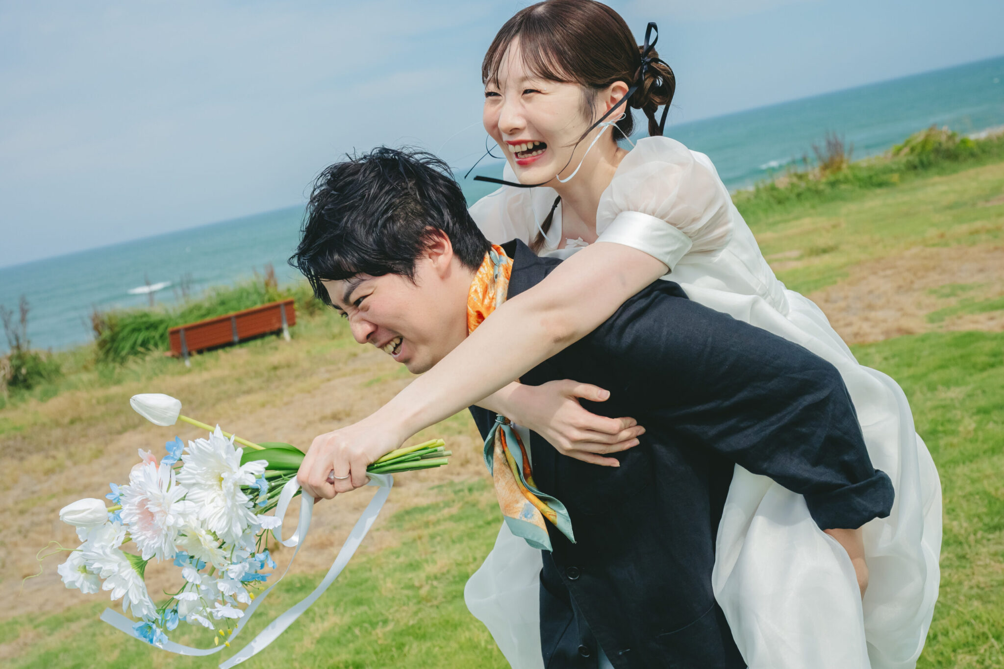 海の見える鹿島灘海浜公園の広い芝生エリアで新婦をおんぶして弾ける笑顔を見せる新郎