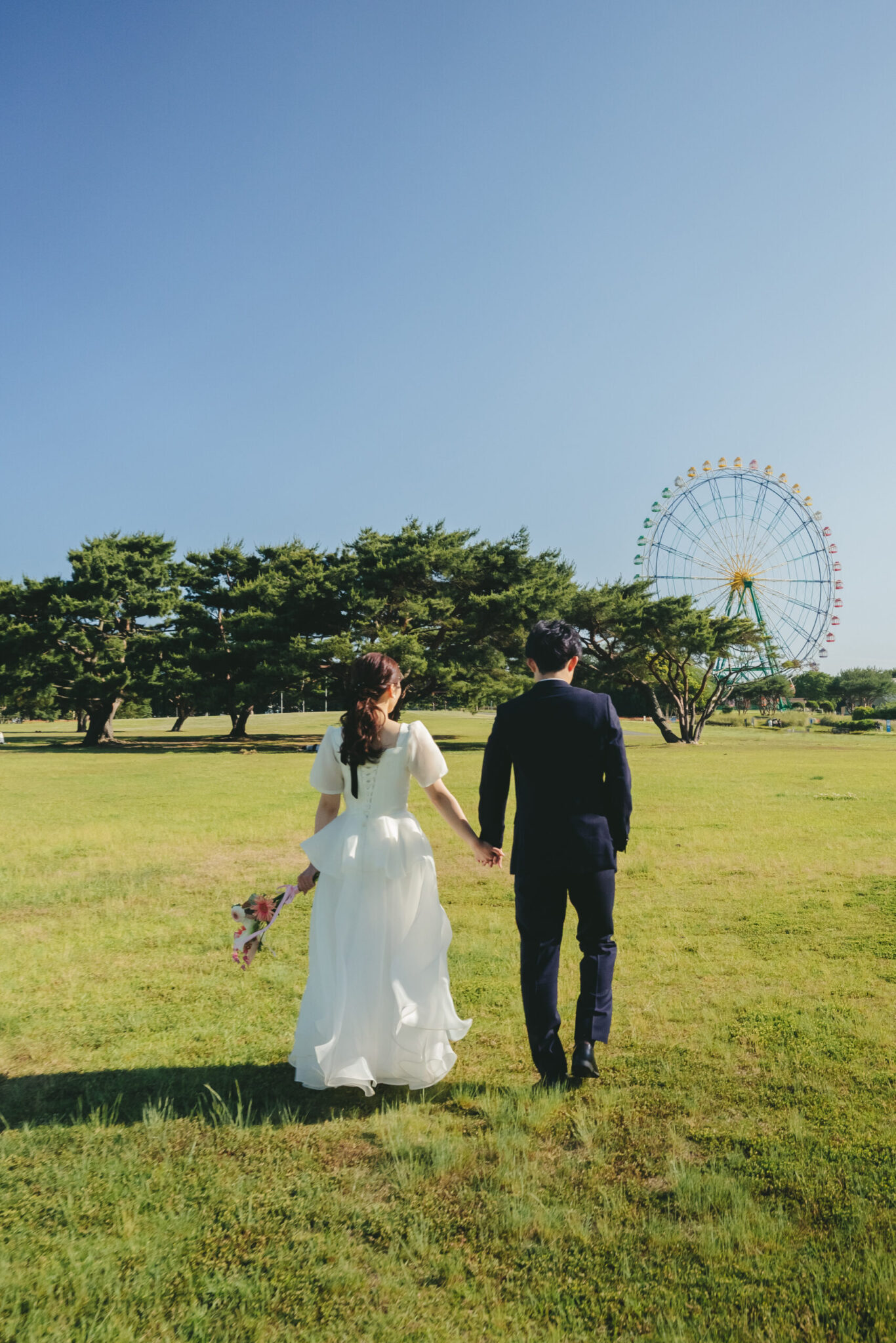夕暮れ時のひたちなか海浜公園で大きな観覧車に向かって手を繋いで歩く新郎新婦の美しい後ろ姿