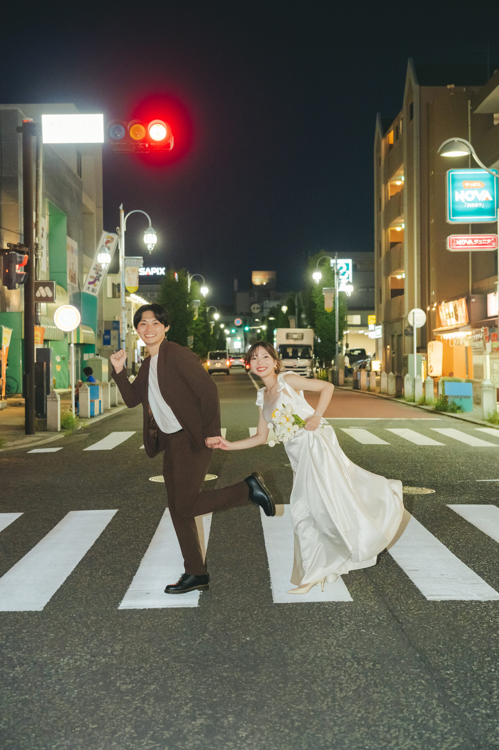 夜の街の信号と横断歩道をドレス姿で手を繋いで渡るスタイリッシュなナイト前撮り