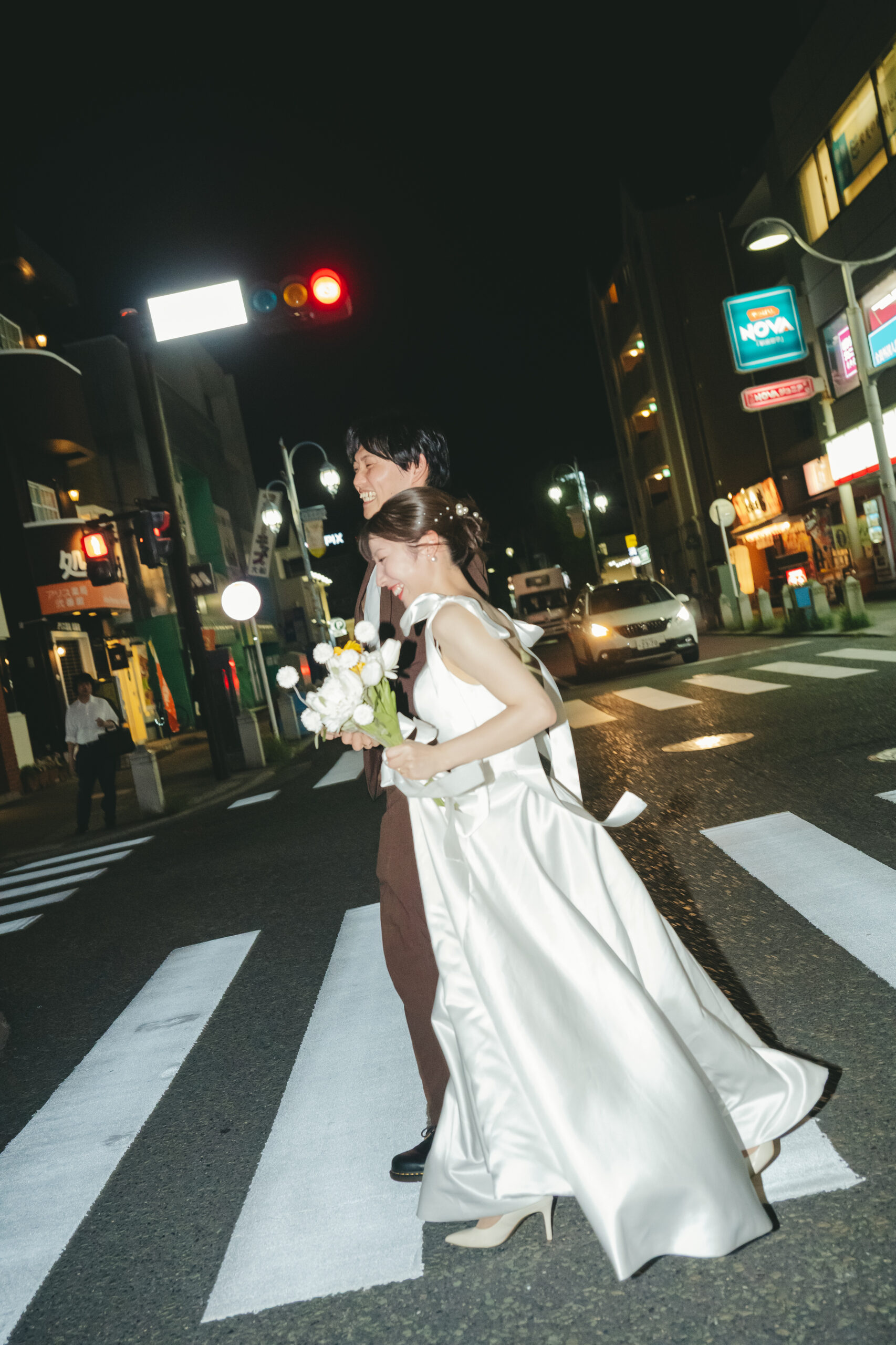 夜の街の信号と横断歩道をドレス姿で手を繋いで渡るスタイリッシュなナイト前撮り
