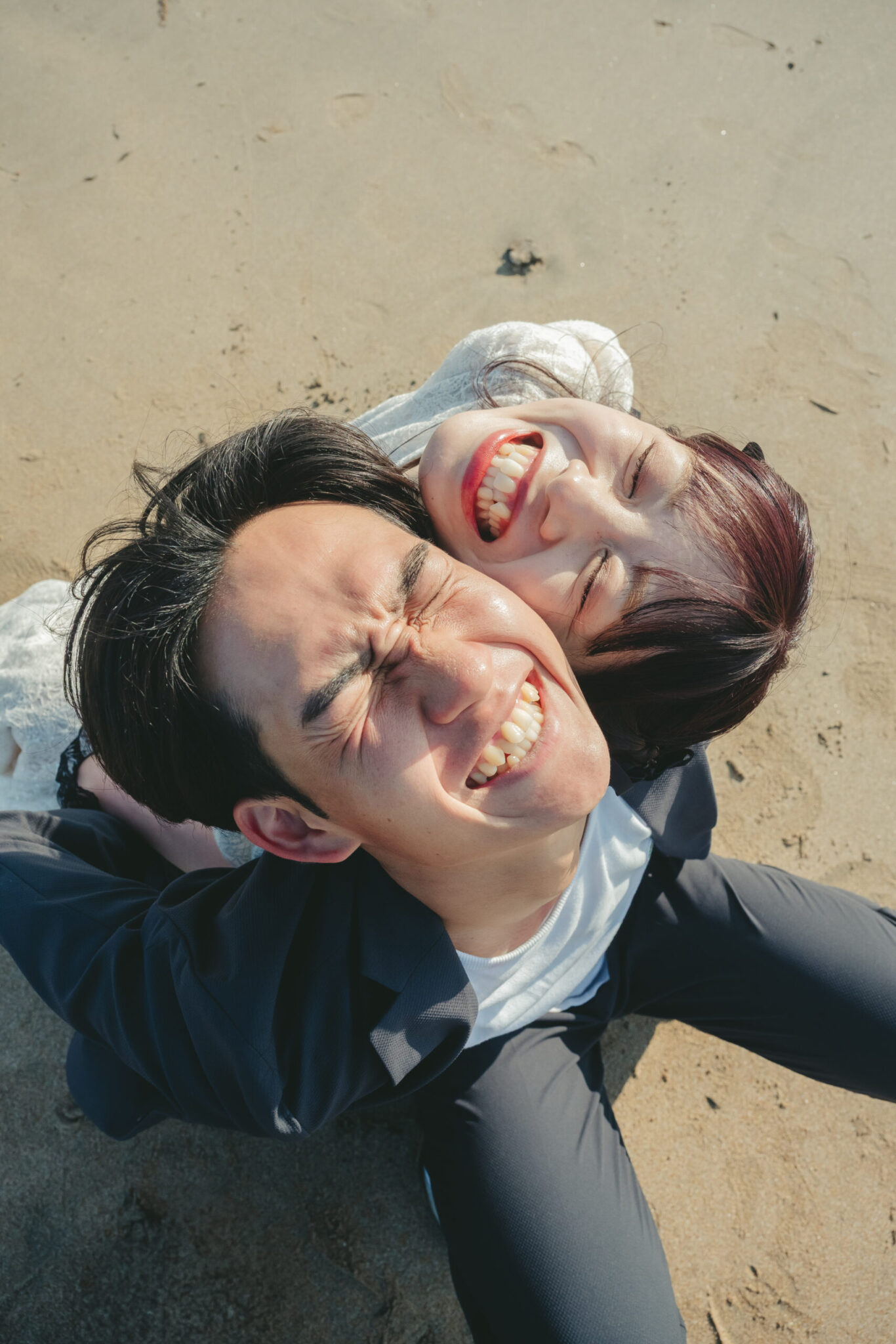 砂浜の上でおんぶをしながらお二人らしい最高に自然体なクシャクシャの笑顔を見せる海前撮りのオフショット