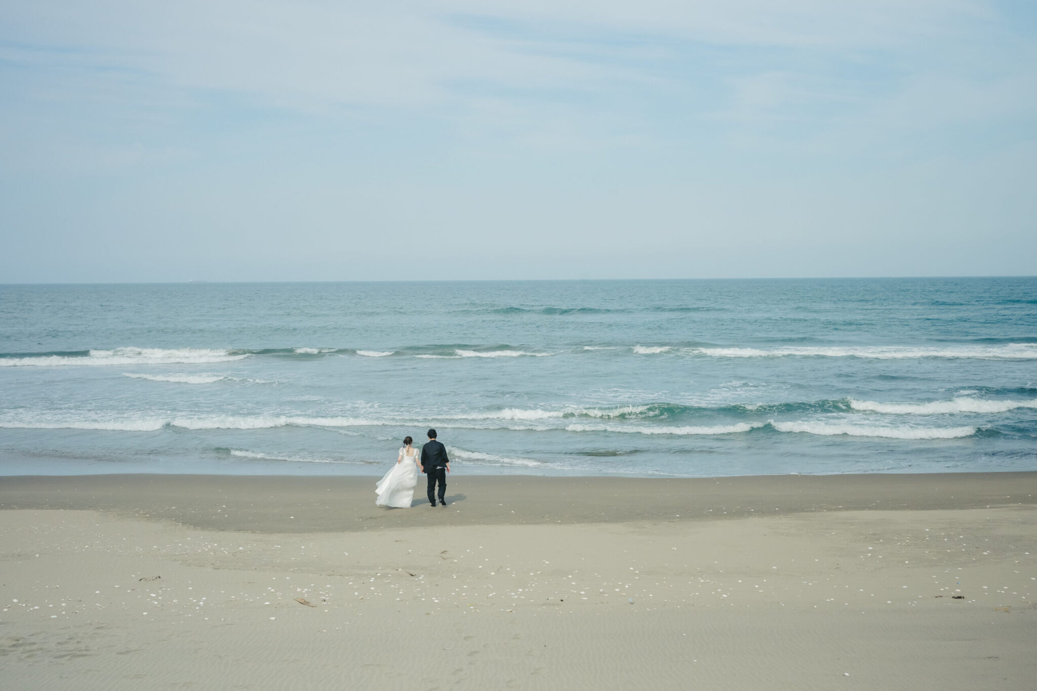 茨城県鹿嶋市の果てしなく広がる砂浜と力強い波の海をバックに映画のように佇む新郎新婦