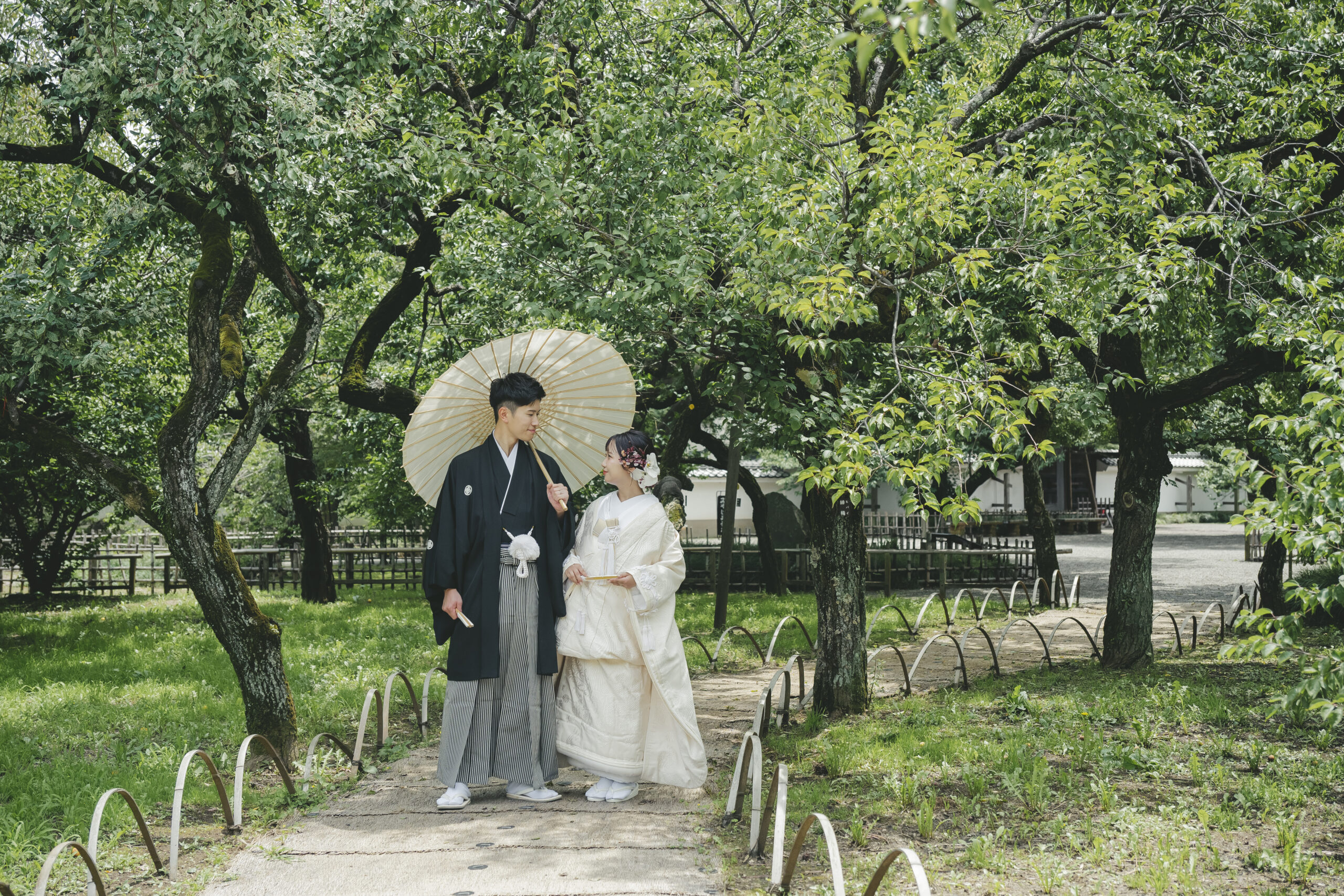 弘道館の豊かな緑に囲まれた庭園の小道を番傘を差して歩く新郎新婦のナチュラルな和装前撮り
