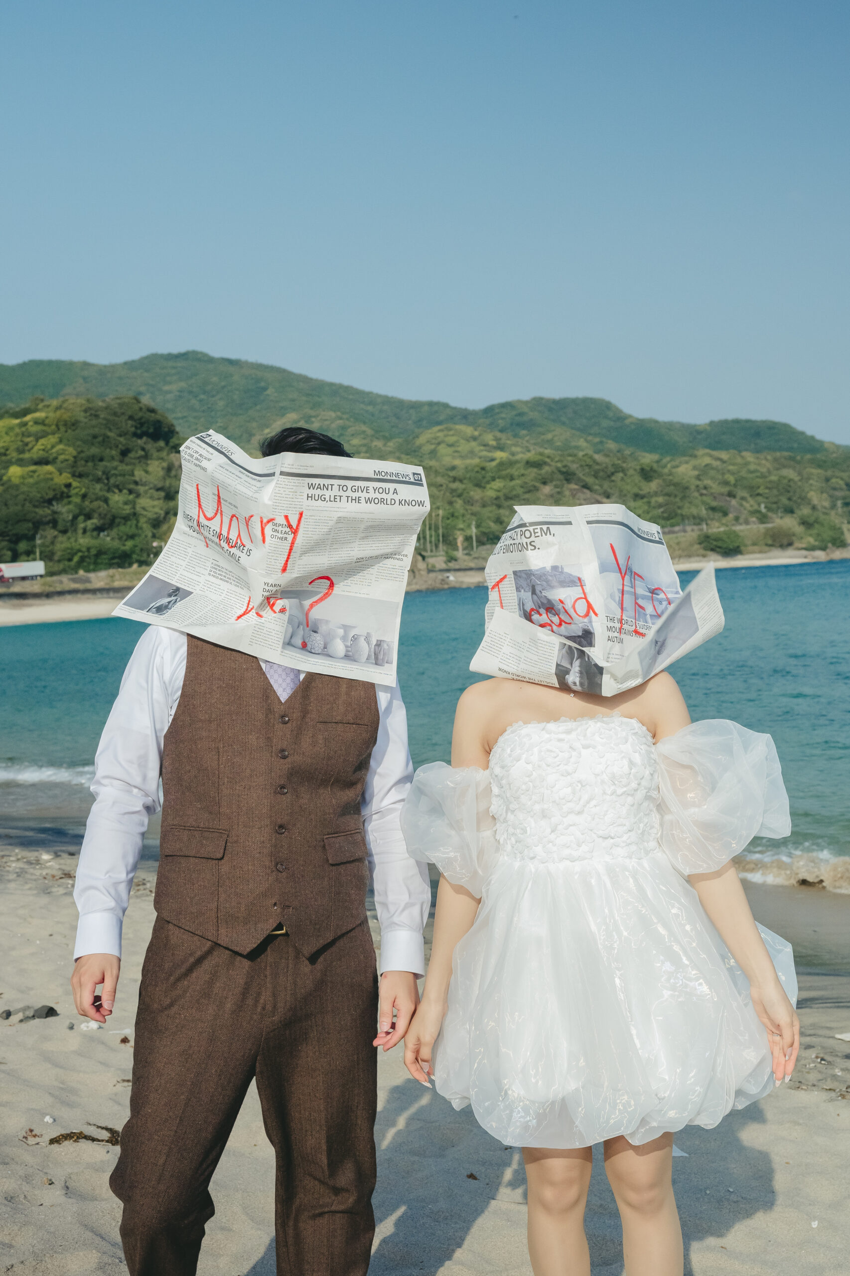 鹿児島のダグリ岬海岸で青い海を背景に英字新聞を持ってポーズを決める前撮りカップル