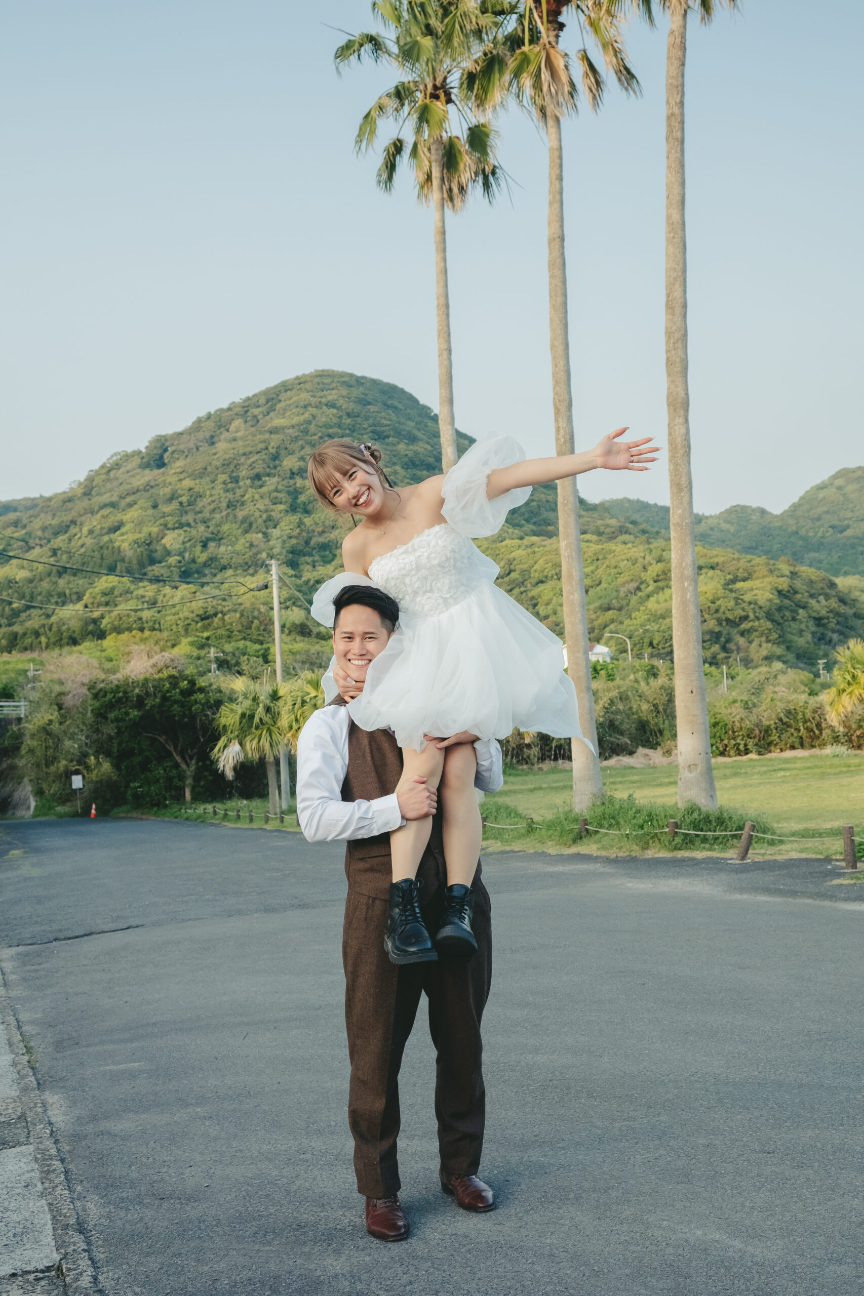 鹿児島のダグリ岬海岸でヤシの木を背景に新婦を抱っこしてポーズを決める前撮りカップル